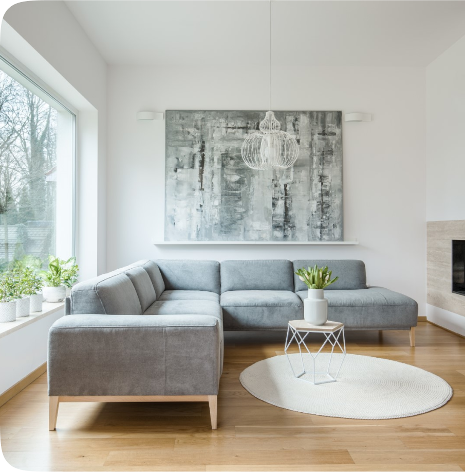 Modern living room with a gray sectional sofa, abstract art, and potted plants. Light decor with hardwood floors and a round, white rug.