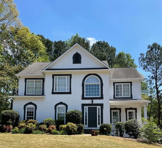 A two-story residential home in Dacula, GA, featuring freshly painted white stucco siding, bold black window trim and molding accents, and a matching black front door illustrating professional exterior painting services.