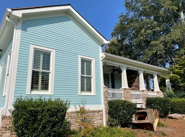 A charming light blue house with white trim, front porch with brick details, and lush greenery. Sunny day with clear sky and trees in the background.