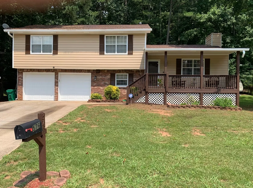 A split-level residential home in Alpharetta, GA, featuring tan horizontal vinyl siding, dark brown window shutters, white garage doors, and a stained wooden front porch deck illustrating professional exterior painting services.