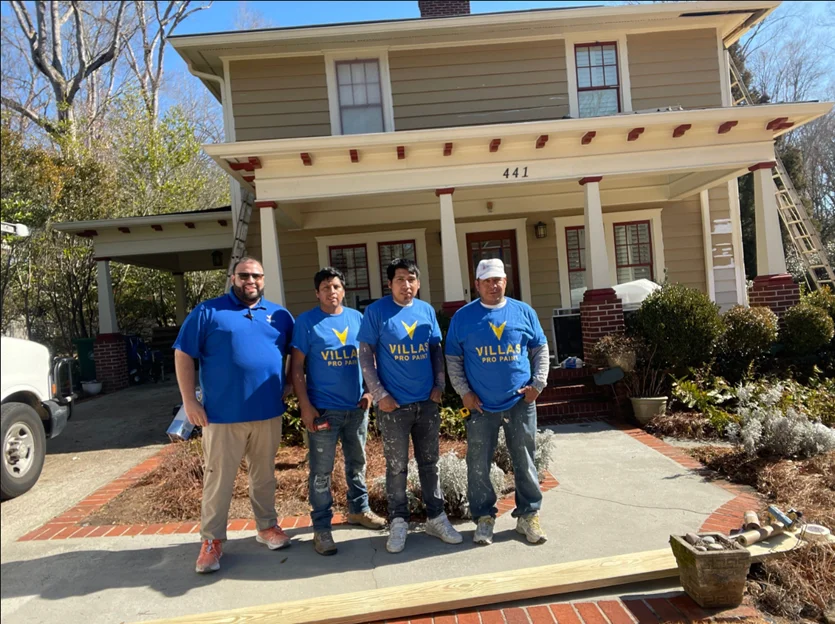 Four smiling men in blue "Villas Pro Paint" shirts stand in front of a freshly painted, beige house with a porch. Sunny day, tools nearby.