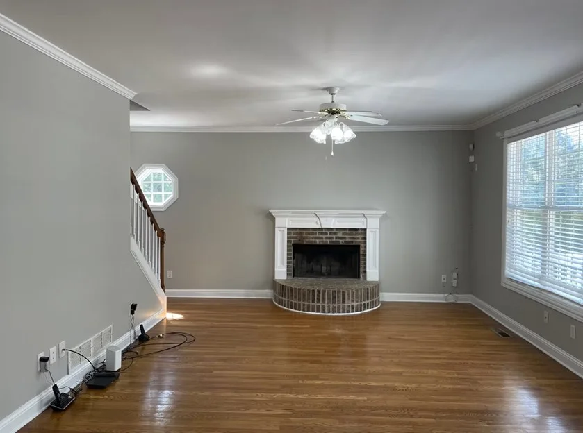 A spacious, open-concept living room in a Dacula, GA home featuring freshly painted light gray walls, crisp white crown molding, a brick fireplace with a white mantel, and polished hardwood floors.