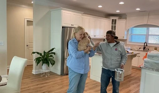 A woman holding a fluffy dog smiles warmly in a cozy kitchen. Beside her, a man with a paint can laughs, creating a joyful and relaxed atmosphere.