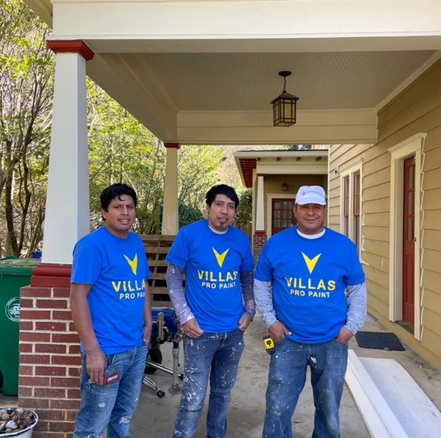 Three painters wearing blue "Villas Pro Paint" T-shirts and jeans stand cheerfully on a porch, conveying teamwork and professionalism.