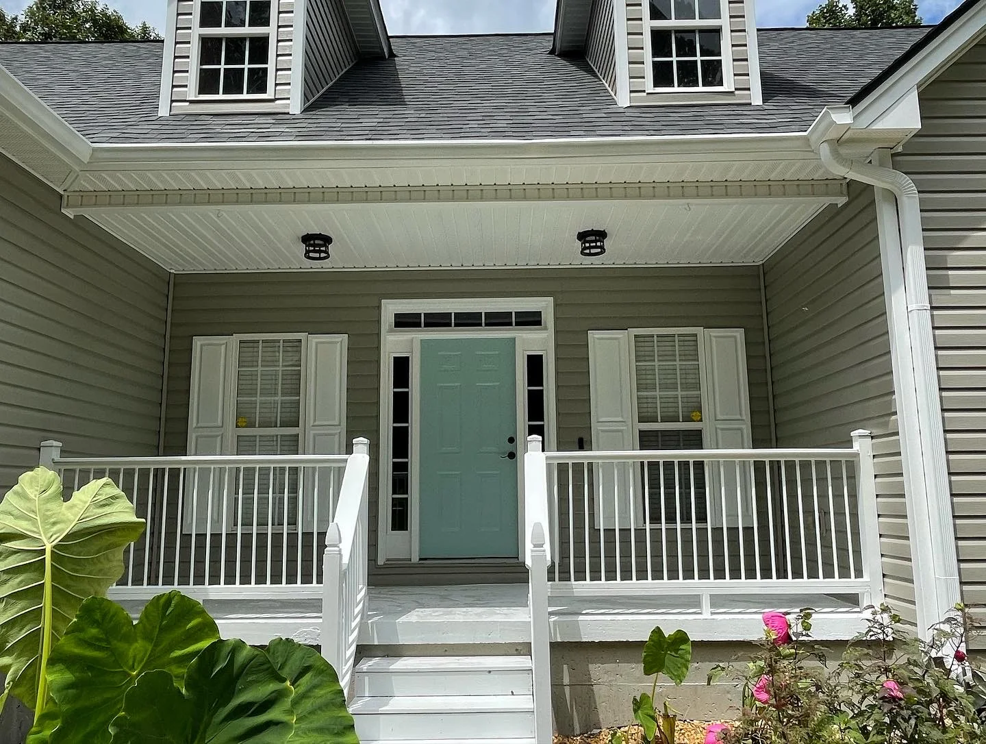 Front porch of a house with light gray siding and a teal door. White railings and steps lead up to it. Vibrant green leaves and pink flowers in the foreground.
