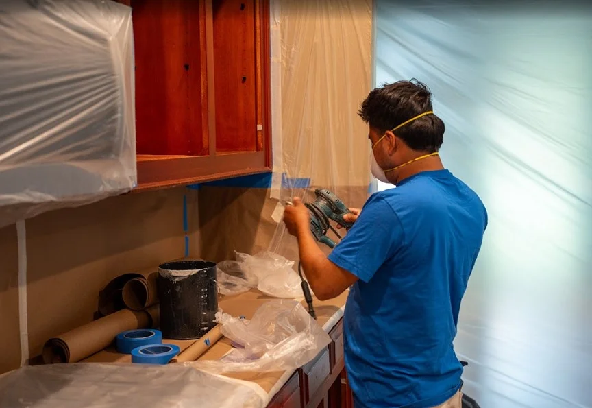 A professional contractor using a power sander to prepare kitchen cabinets for painting, illustrating the detailed surface preparation and labor that influence interior painting costs in Alpharetta, GA.