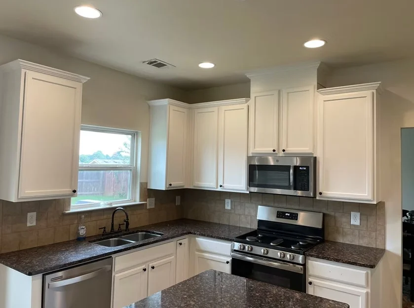 A modern kitchen in a Dacula, GA home featuring freshly painted white cabinets, dark granite countertops, stainless steel appliances, and matching white window trim.
