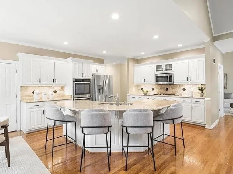 A large, modern kitchen in a Dacula, GA home featuring freshly painted white cabinets, a matching kitchen island with gray bar stools, neutral beige walls, and polished hardwood floors.