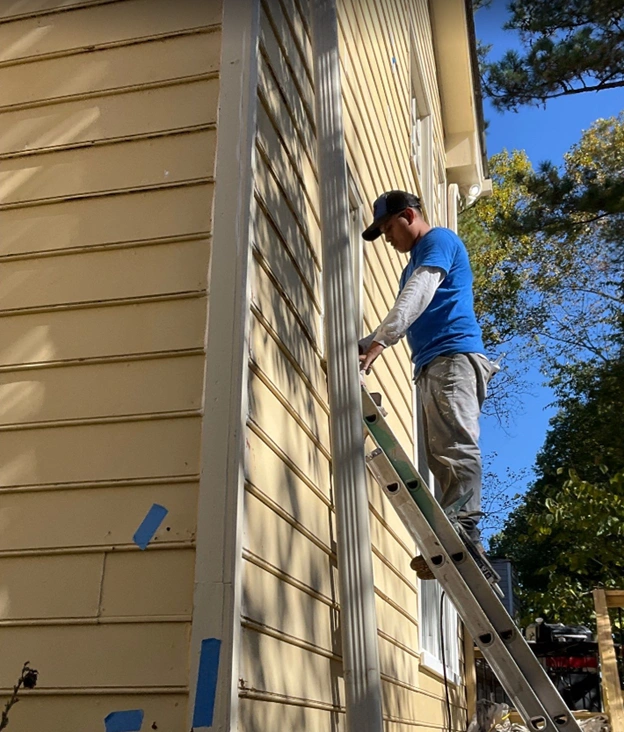 A professional contractor standing on an extension ladder while performing meticulous siding preparation on a yellow Alpharetta home, demonstrating how exterior painting protects house materials from humidity and weather damage.