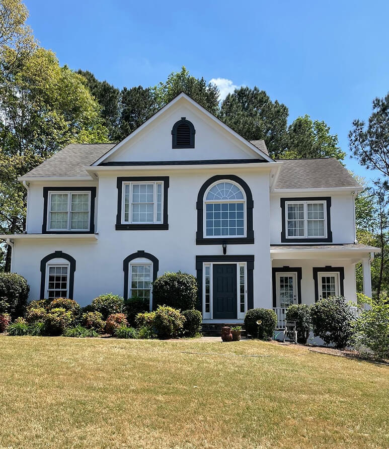 Colonial-style house with white walls, dark trim, and large arched window. Surrounded by green shrubs, under a clear blue sky, conveying a serene ambiance. Local House Painters