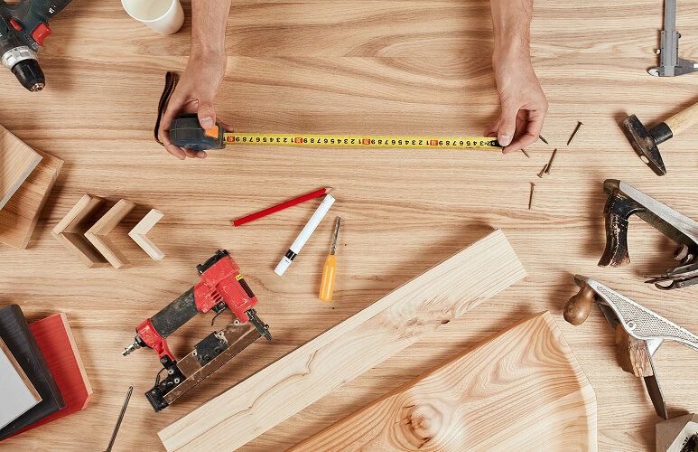 Overhead view of a woodworking workspace with tools. Hands hold a measuring tape across a wooden table, surrounded by a drill, nails, saw, and wooden planks.