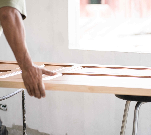 A person fits a wooden frame with multiple sections near a bright window, suggesting crafting or assembly, creating a focused and calm atmosphere.