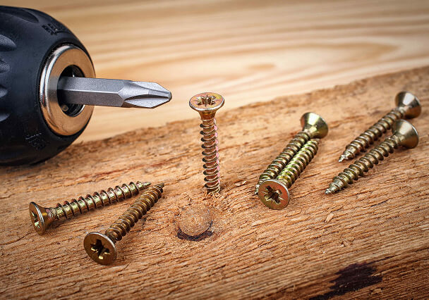 A black screwdriver is positioned near several brass screws on a wooden surface. One screw is partially inserted, indicating active work in progress.