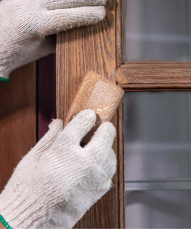 Hands in gray gloves sanding a wooden window frame, emphasizing the texture and grain. The setting suggests a careful, detailed restoration task.