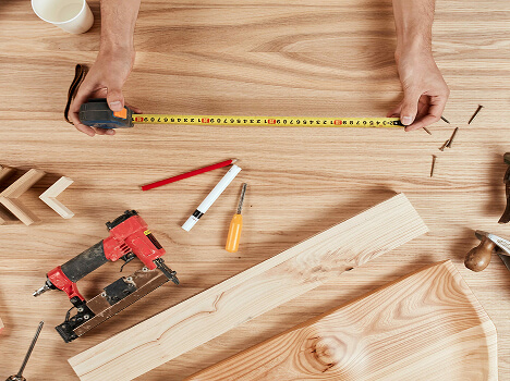 Hands measure wood with a tape measure on a workbench surrounded by carpentry tools, pencils, and screws, conveying a DIY woodworking project.
