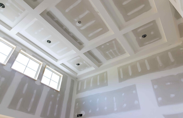 High ceiling with recessed lighting in a large room under construction. Unfinished drywall panels show seams and patches, and sunlight streams in from three windows.