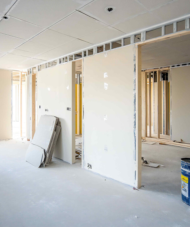 A bright, unfinished room with drywall sheets stacked against a wall, exposed wooden framing, and a blue paint can on the floor, suggesting ongoing construction.
