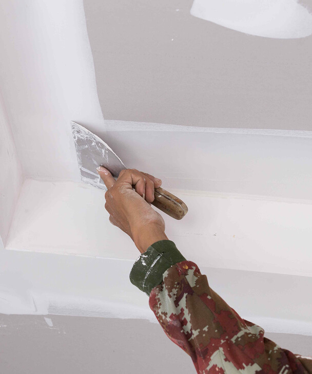 A person in a camouflage jacket smoothly applies white plaster on a wall corner using a putty knife, showcasing focus and craftsmanship.