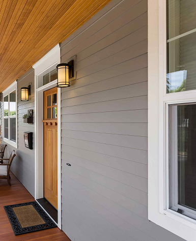 Elegant porch with a wooden door, framed by two modern lanterns. The gray siding contrasts with the ceiling's warm wood tones, creating a welcoming entryway.