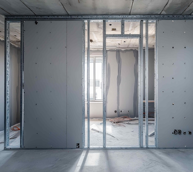 Construction scene with unfinished partition walls using metal studs and drywall in a sunlit room. Exposed wires and light through the window.