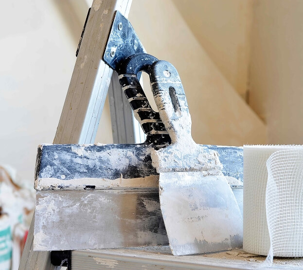 A plaster-covered plastering trowel rests on a ladder in a home renovation setting. Nearby, mesh tape sits on a surface, evoking a sense of ongoing work.