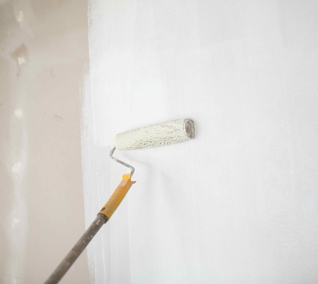 A paint roller applies white paint on a wall, creating a fresh, clean surface. The handle is yellow, and the roller moves smoothly, indicating progress.