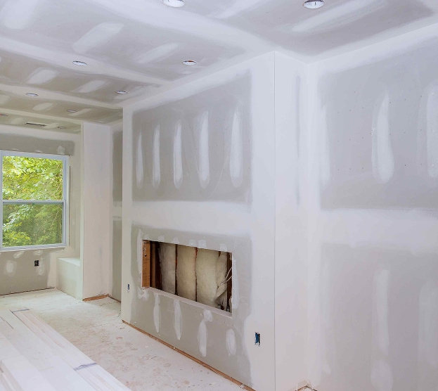 Unfinished room with drywall, recessed lights, and window showing greenery. Wall cut-outs and fresh plaster indicate ongoing construction work.