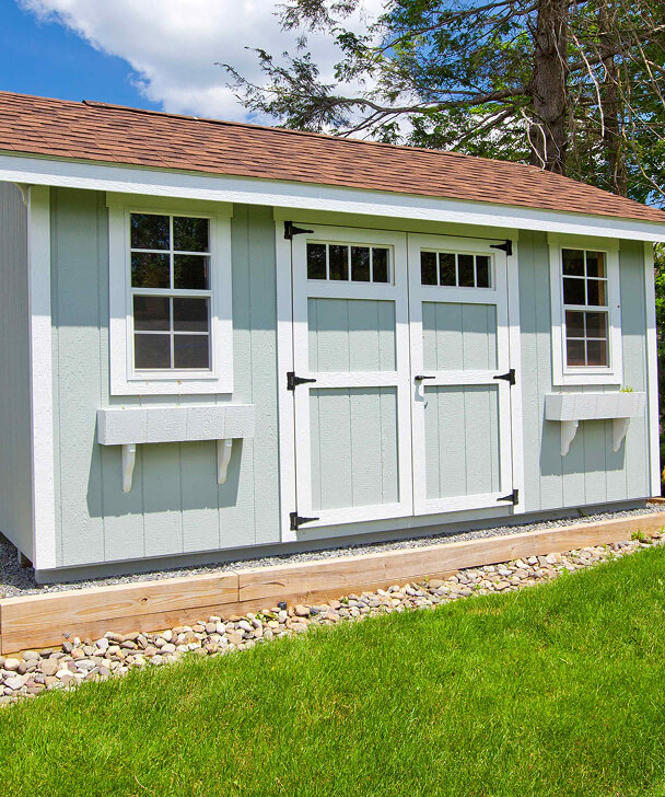 A quaint light gray garden shed with white trim, brown shingled roof, and double doors, surrounded by lush green grass and a gravel border.