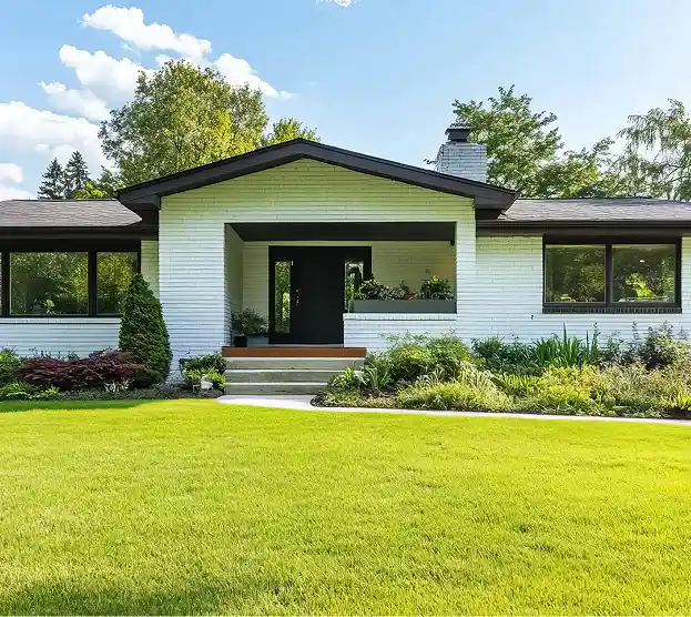 A modern, single-story white brick house with a dark roof, surrounded by lush greenery and a well-manicured lawn, under a clear blue sky.