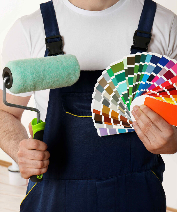 Man in navy overalls holds a paint roller and a color palette fan. The scene suggests home improvement, conveying preparedness and creativity.