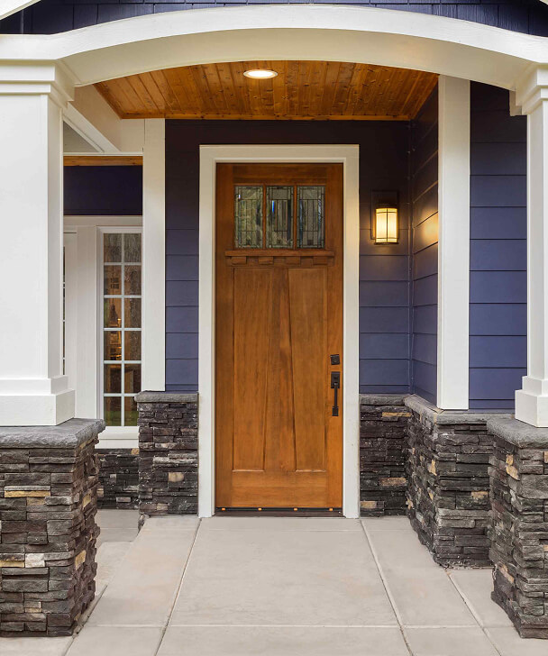 Wooden front door with glass panels, surrounded by blue siding and stone accents. A warm porch light gives a welcoming feel to the entrance.