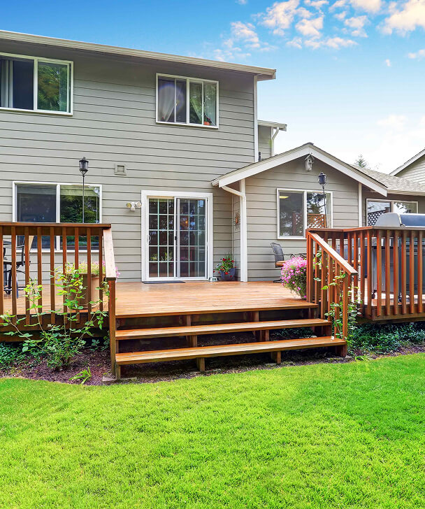 Two-story gray house with a wooden deck featuring steps, railings, and potted pink flowers. Vibrant green lawn under a blue sky with light clouds.