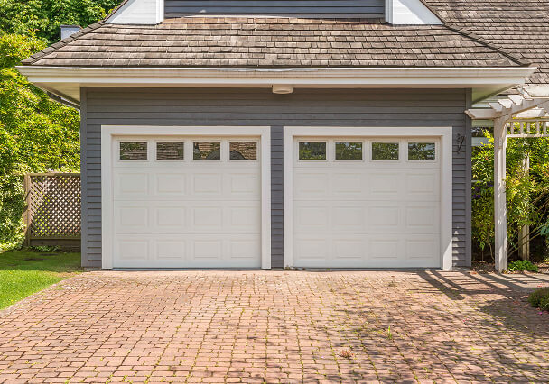 A double-garage with white doors and small windows sits against a blue house. The driveway is paved with red bricks, surrounded by green shrubs.