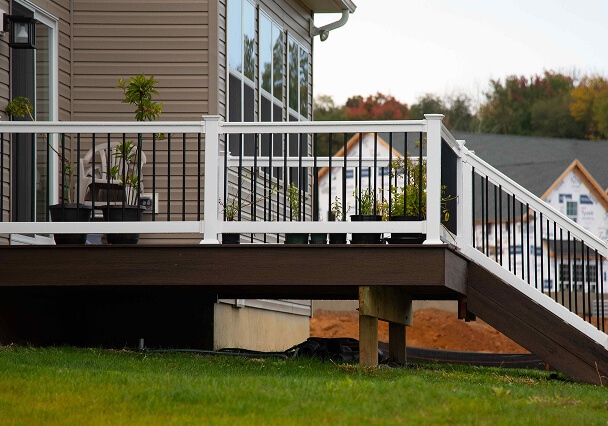 A wooden deck with white railing is adorned with potted plants. It connects to a house with large windows. Houses under construction are in the background.