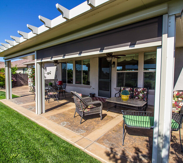 A sunny patio with a pergola features wicker chairs with colorful cushions, a coffee table, and potted plants. The scene conveys warmth and relaxation.