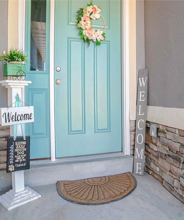 Light blue front door with a floral wreath, flanked by a tall "WELCOME" sign. A welcome stand with potted plant and doormat adds a cozy greeting.