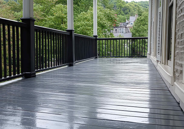 Wide porch with glossy dark gray wooden floor, surrounded by a black railing. Lush green trees and rooftops visible in the background, evoking serenity.