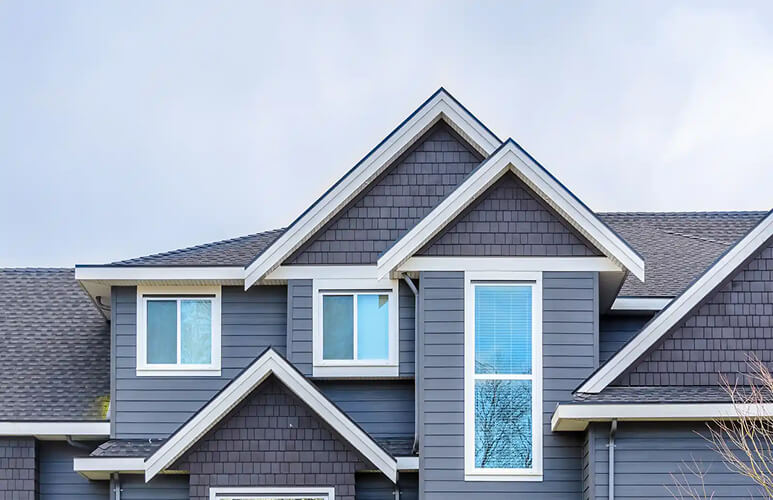 Gray modern house exterior showcasing multiple peaked roofs, white-trimmed windows, and a cloudy sky background, conveying a sleek, contemporary feel.