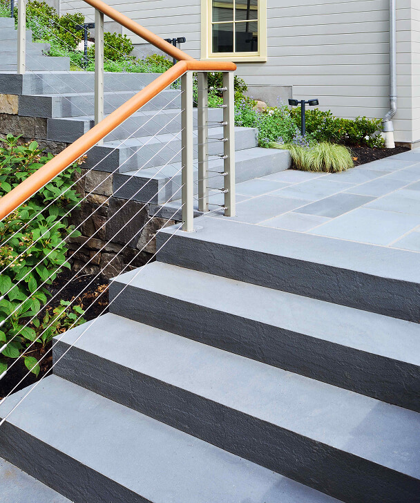 Wide stone steps with a sleek metal and wood handrail lead up to a modern house. Green shrubs line the path, adding a touch of nature.