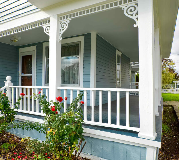 A charming porch with light blue siding, white trim, and ornate accents. Red roses bloom in front, adding a vibrant touch to the serene scene.