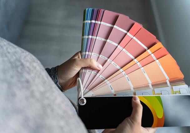 A person holds a fanned-out color swatch book displaying various shades from pink to orange. The setting is well-lit, conveying a sense of creativity.