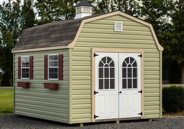 A small, light green shed with a brown shingled gambrel roof, double white doors, and decorative arched windows. Red shutters add a rustic touch.