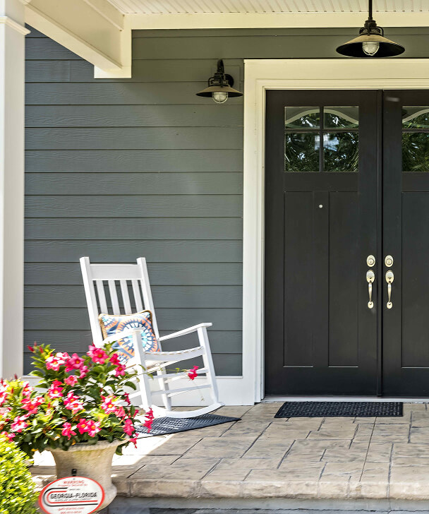 Front porch with a white rocking chair and colorful cushion beside a vibrant potted plant. Black double doors and serene gray siding create a welcoming feel.