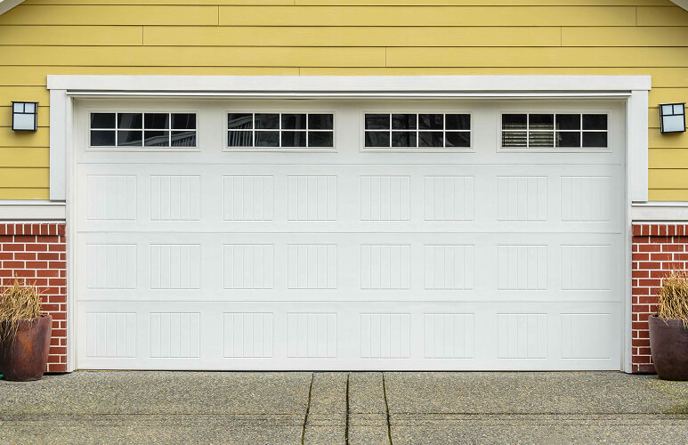 White garage door with rectangular windows at the top, set against a yellow wall and red brick base. Flanked by two black wall lanterns, conveying a tidy, suburban feel.