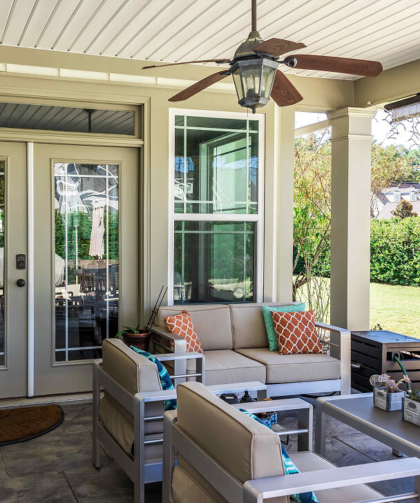 Cozy patio with beige seating, colorful pillows, and glass-topped tables. Ceiling fan above; double doors open to a green, sunlit backyard. Relaxed vibe.