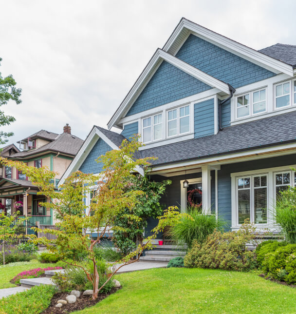 Charming two-story blue house with white trim, surrounded by lush greenery and a manicured lawn, exudes a welcoming and serene suburban feel.