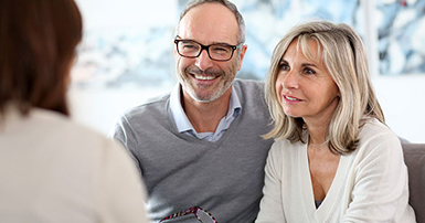 A smiling middle-aged couple sits on a couch, attentively listening to a person in front of them. The setting is warm and cozy, conveying an atmosphere of comfort.