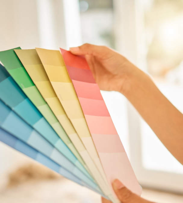 A hand holds a fan of color swatches in a gradient from blue to pink. The background is softly blurred, suggesting an interior setting with natural light.