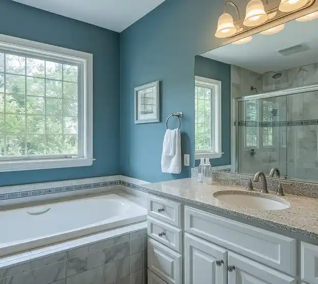 A serene bathroom with blue walls features a large bathtub near a window, a white vanity with a granite countertop, and an elegant light fixture.