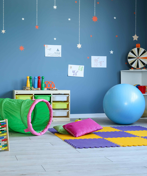 Bright playroom with a blue wall, colorful foam mat, green play tunnel, exercise ball, and shelves of toys. Star decals and kids' drawings adorn the wall.